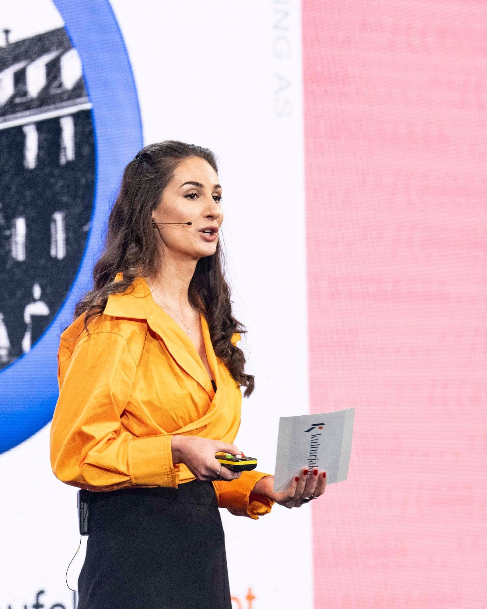 Hana Colakovic on stage in an orange blouse, holding Kulturjakten cards